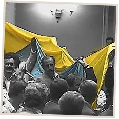 A crowd indoors raising a large Ukrainian flag overhead.